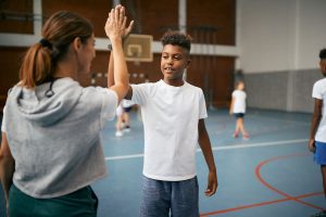 Happy black elementary student and his sports teacher giving high-five during PE class at school gym.